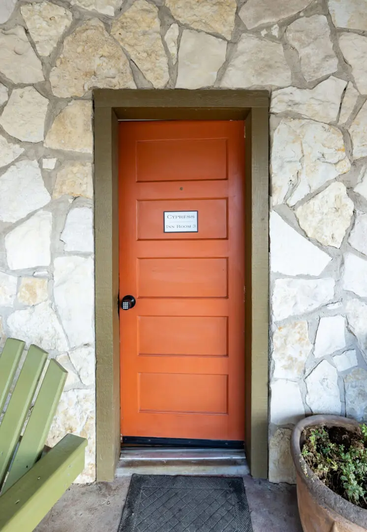 A orange door on a wall with a stone design with flower pot kept on the side