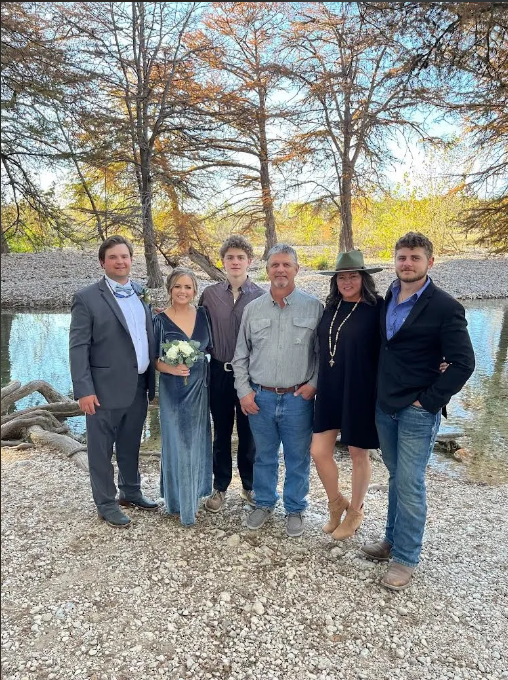 A group of six people stands outdoors by a river, posing for a photo. Trees and rocky terrain surround them, capturing the natural beauty of Leakey Texas. Perfect Lodging near the Frio River can be seen in the background.