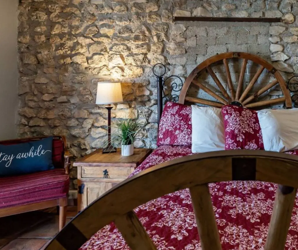 A cozy bedroom with a stone wall, a bed with a red floral comforter, a wooden nightstand holding a lamp and a plant, and a cushioned chair with a "stay awhile" pillow. Experience charming Leakey Texas lodging near the Frio River for your next relaxing getaway.