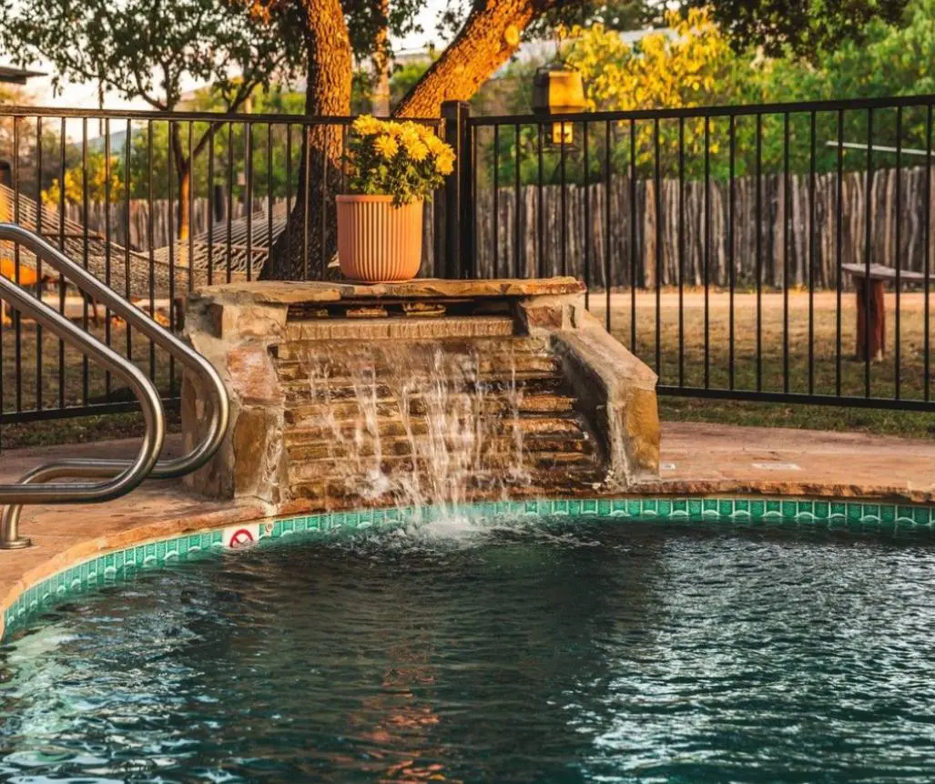 Experience a serene swimming pool with a captivating waterfall feature, surrounded by a black fence and lush trees at Leakey Texas Lodging near the Frio River. A potted plant sits elegantly on the stone structure above the waterfall, while a hammock invites relaxation in the background.